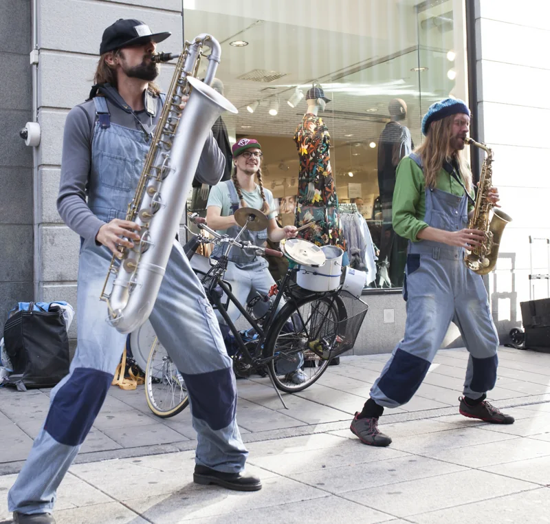 Two saxophonists in dungarees dancing down a street with the drum bicycle behind them.